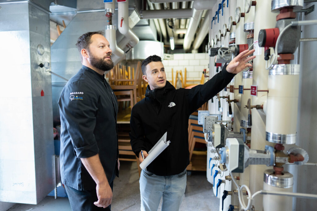 zwei Männer stehen im Heizungsraum und betrachten Leitungen